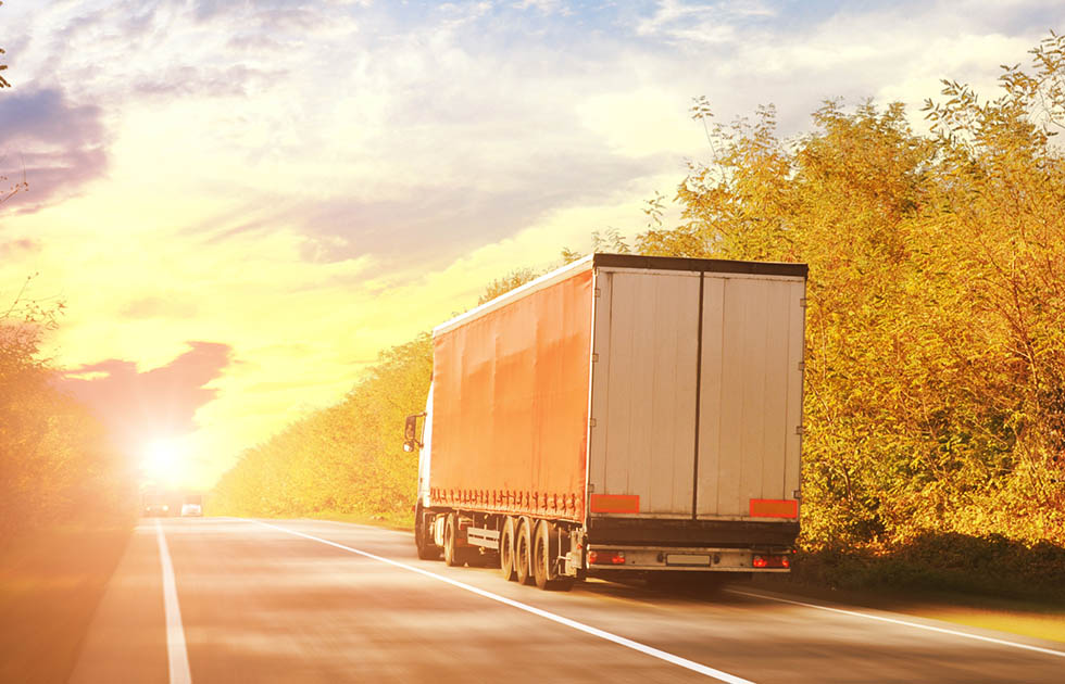 A truck drives along a road, silhouetted against a vibrant sunset sky filled with orange and purple hues